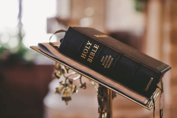 A close-up photograph of a black leather-bound Holy Bible with gold lettering, resting on a golden stand.