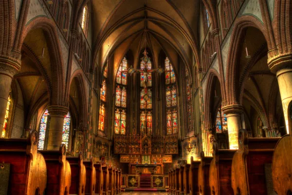 The interior of a grand church with rows of wooden pews leading to a vibrant altar bathed in light from large, ornate stained glass windows.