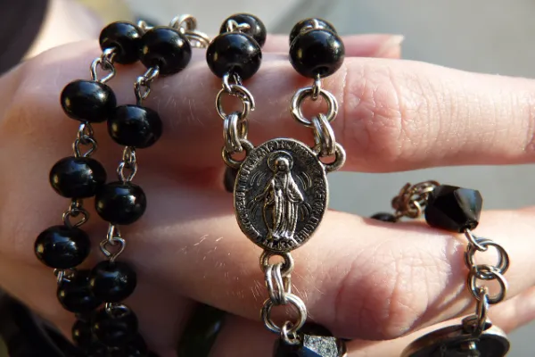 A close-up of a rosary with shiny black beads and a silver medal featuring a depiction of the Virgin Mary, held in a person's hand.