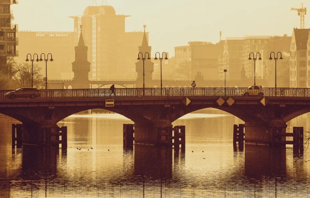 Berlin's Oberbaum Bridge, featuring two historic brick towers, arches over the Spree River at sunset, with a person walking on the walkway and modern city buildings silhouetted in the warm, hazy background.