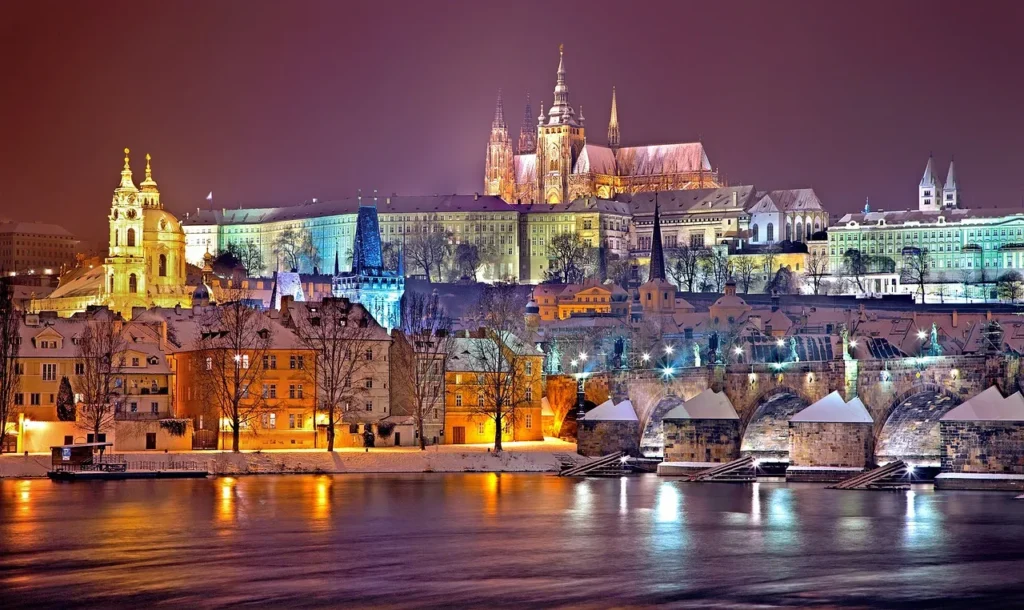 The illuminated nighttime skyline of Prague, Czech Republic, featuring the historic Charles Bridge in the foreground, and Prague Castle and St. Vitus Cathedral dramatically lit on the hill overlooking the Vltava River.