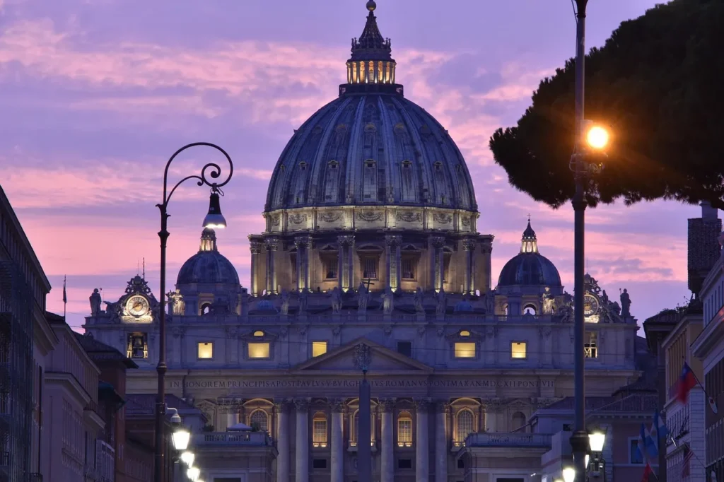 St. Peter's Basilica in Vatican City, with its grand dome and classical facade illuminated at dusk against a vibrant purple and pink sky, framed by streetlights and trees.