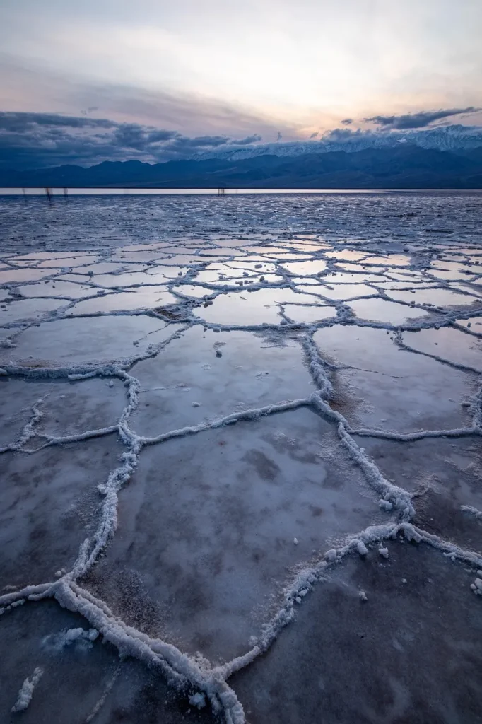 A desolate landscape of hexagonal salt crystal formations on a flat valley floor, reflecting a cold blue sunset or twilight sky, with snow-capped mountains visible in the distance, symbolizing extreme, cold, and challenging travel destinations.