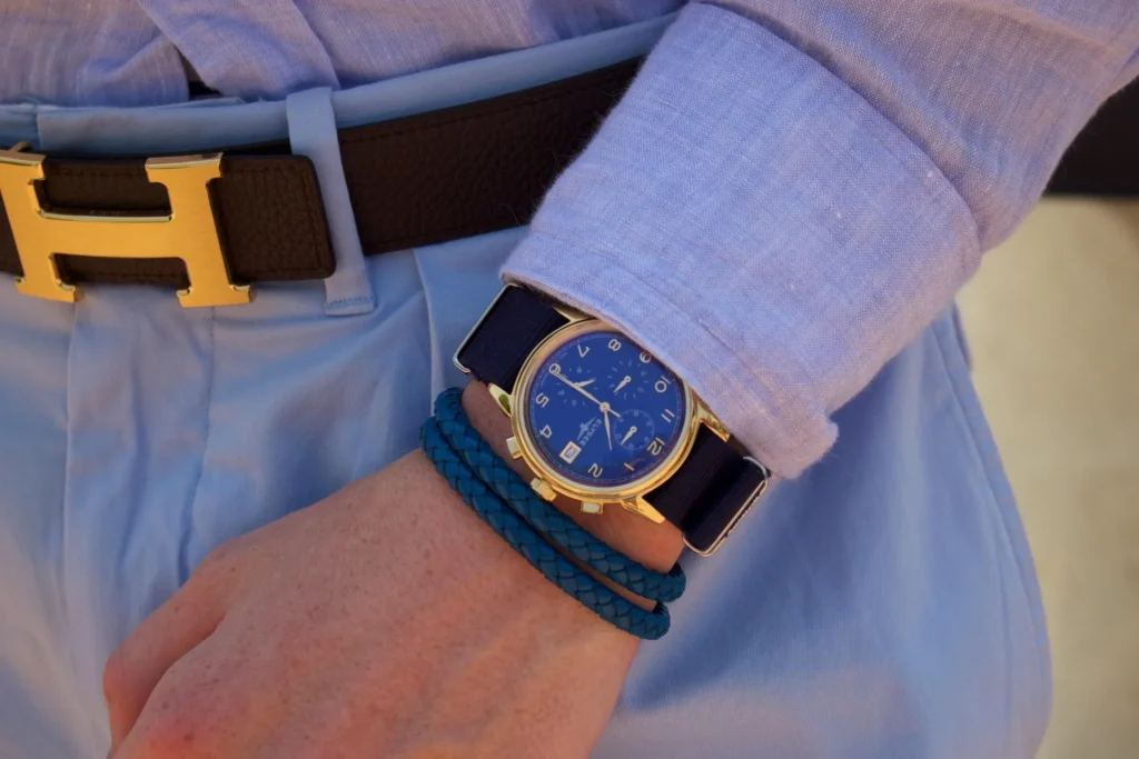 A close-up of a person wearing luxury accessories, including a gold-tone Elysee chronograph watch on a navy NATO strap, a blue braided leather bracelet, and a brown leather belt with a prominent gold ‘H’ buckle. The ensemble is paired with a light blue linen shirt and trousers, symbolizing a high-end, curated lifestyle.